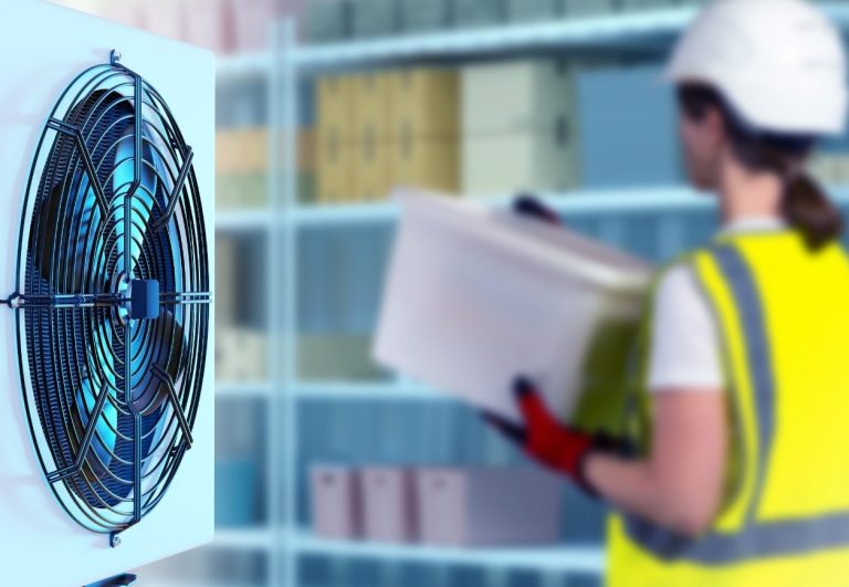 A worker in a hard hat and safety vest examines a book near a large evaporator fan inside a cool room.