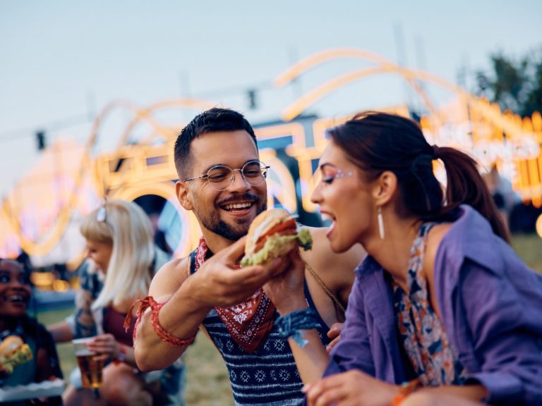 A man offers a large burger to a woman at a lively outdoor festival.